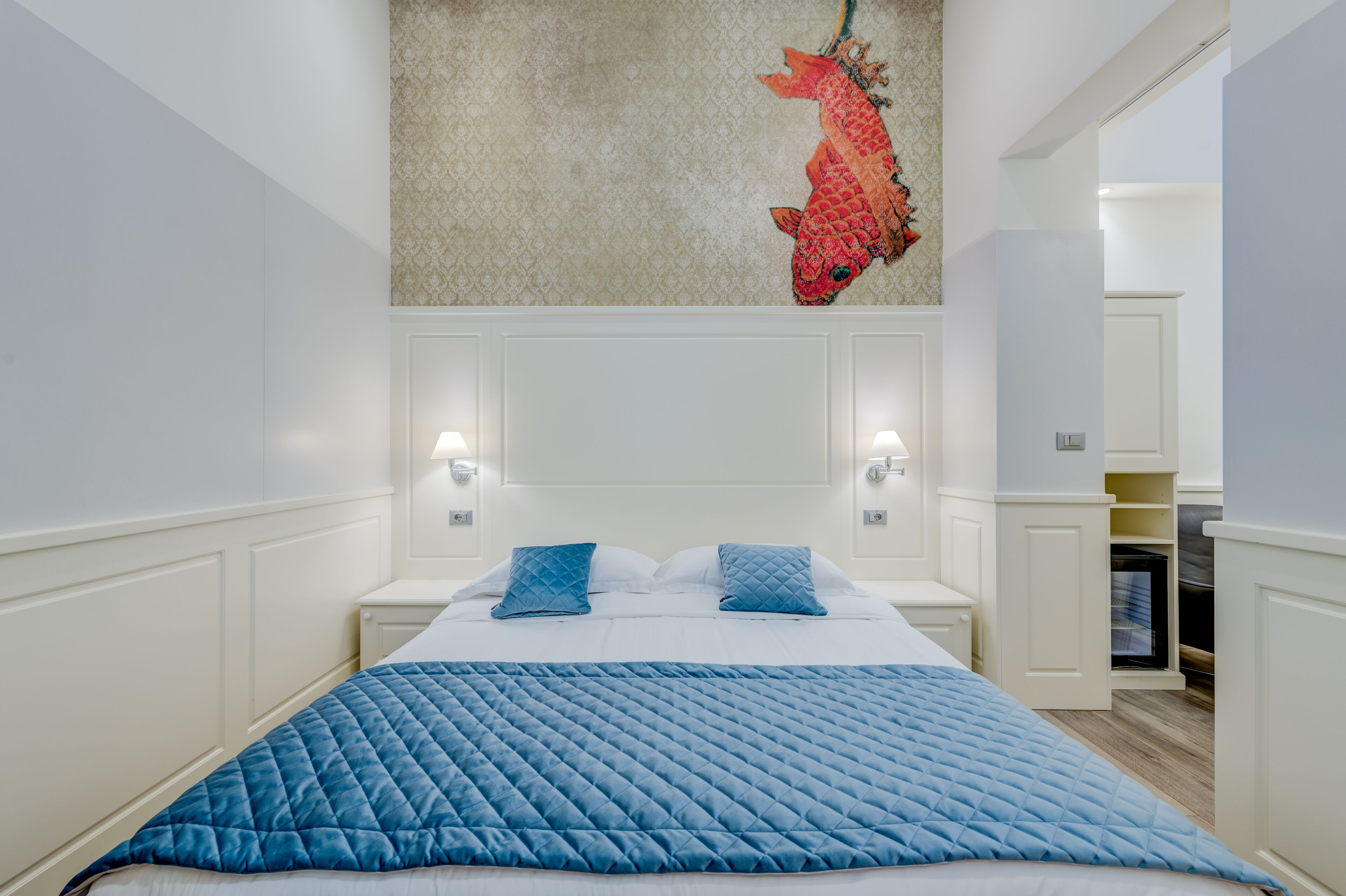 A bedroom with a bed, blue bedding, a decorative fish wall hanging, and a view of a kitchen area.