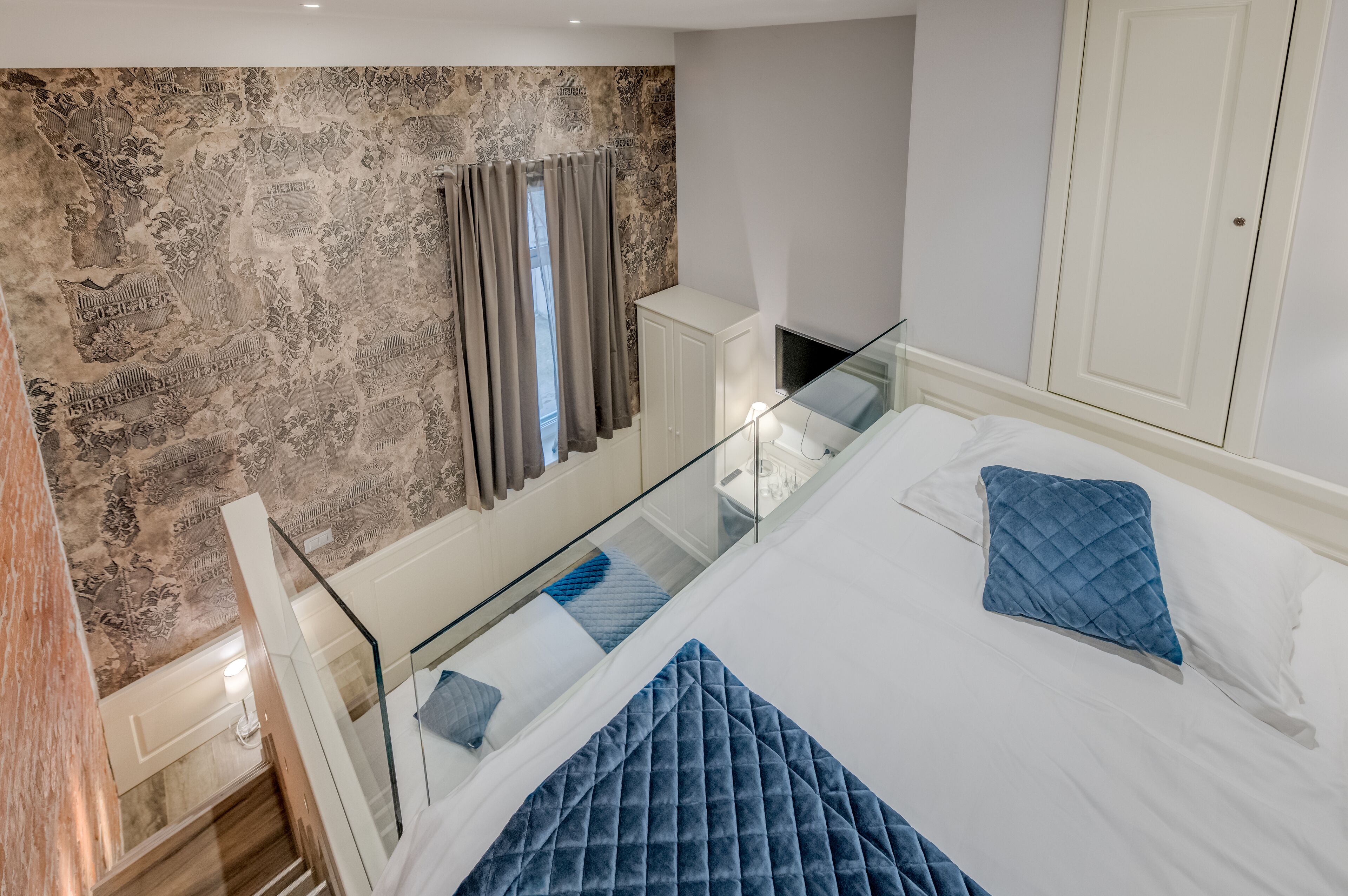 A bedroom with a bed, a glass railing, a wardrobe, and a patterned wall.