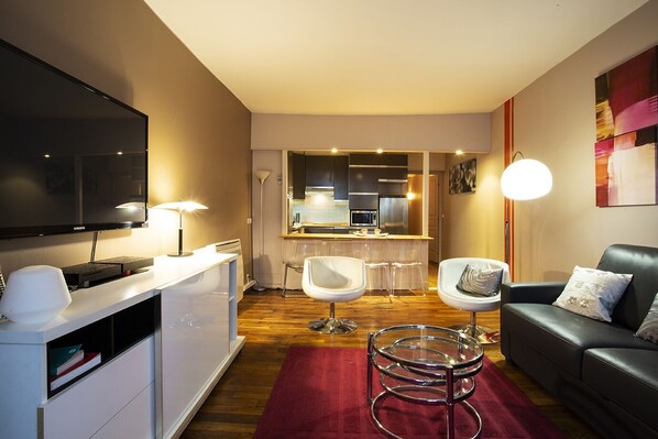 A modern living room with a flat-screen TV, a white desk, a red rug, a black leather sofa, and a glass coffee table.