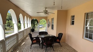 Une terrasse couverte avec une table ronde et des chaises, un ventilateur de plafond et une vue sur la verdure environnante.