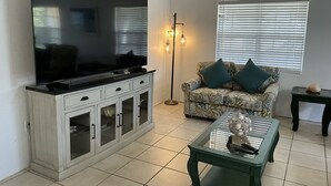 Un salon avec un téléviseur à écran plat posé sur un meuble, un canapé à motifs agrémenté de coussins, une table basse ornée d’objets décoratifs et un lampadaire.