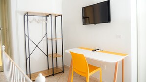 A room with a white desk, a yellow chair, a black TV mounted on the wall, and a metal shelving unit with clothes hanging. - Vulcania Rooms (Naples)
