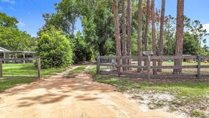 Un chemin de terre mène à une zone délimitée par une grille, avec une maison qui se profile à l’arrière-plan.