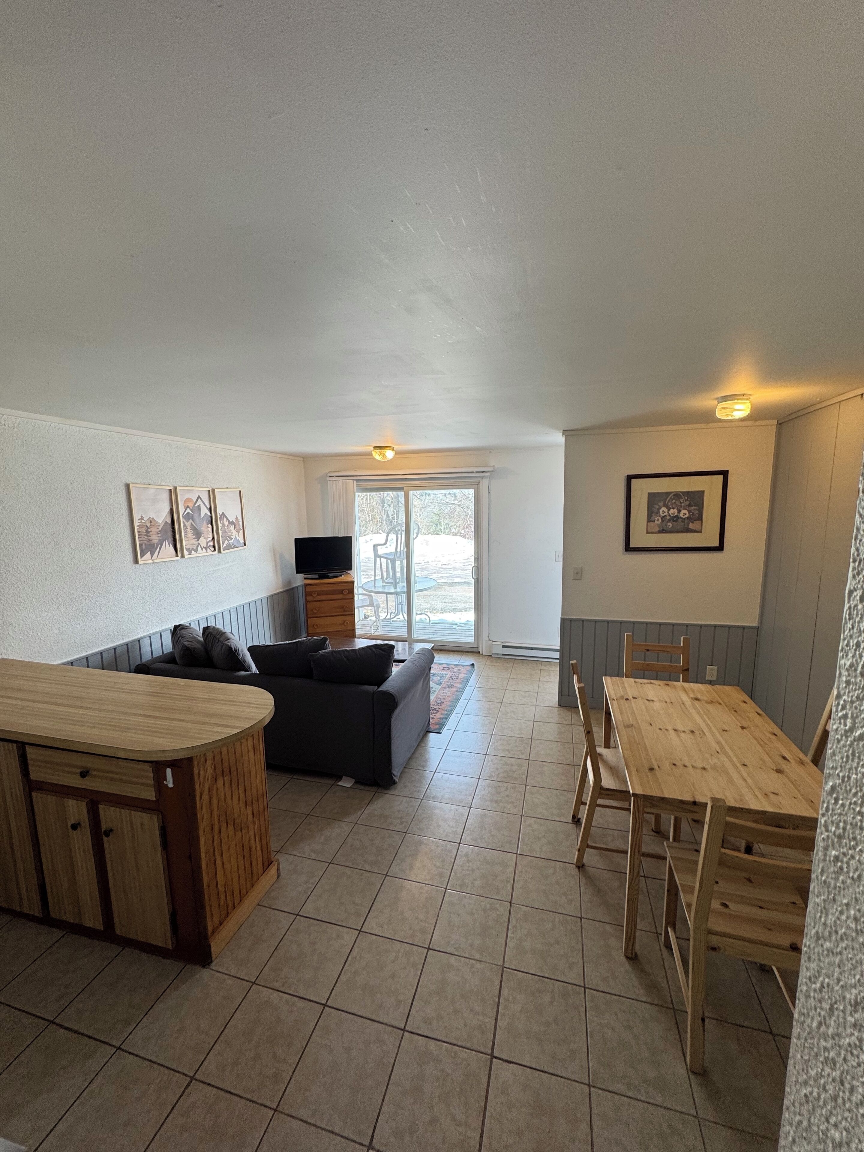 A spacious living area with a sofa, a TV, and a dining table with chairs. There is a sliding glass door leading to an outdoor area.