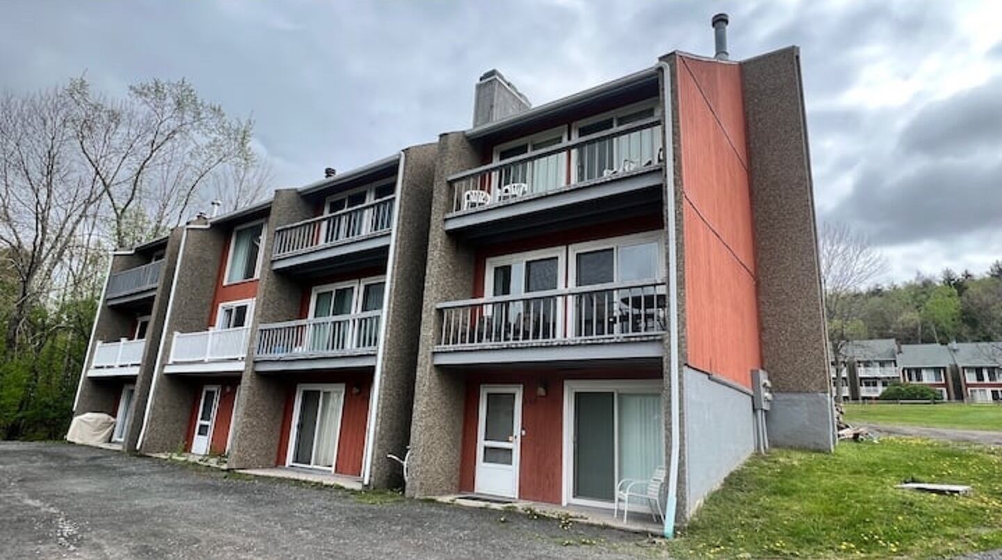 A multi-story apartment building with balconies, a parking area, and a grassy landscape.