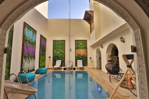 An indoor swimming pool with clear glass walls, surrounded by lounge chairs and a decorative archway.