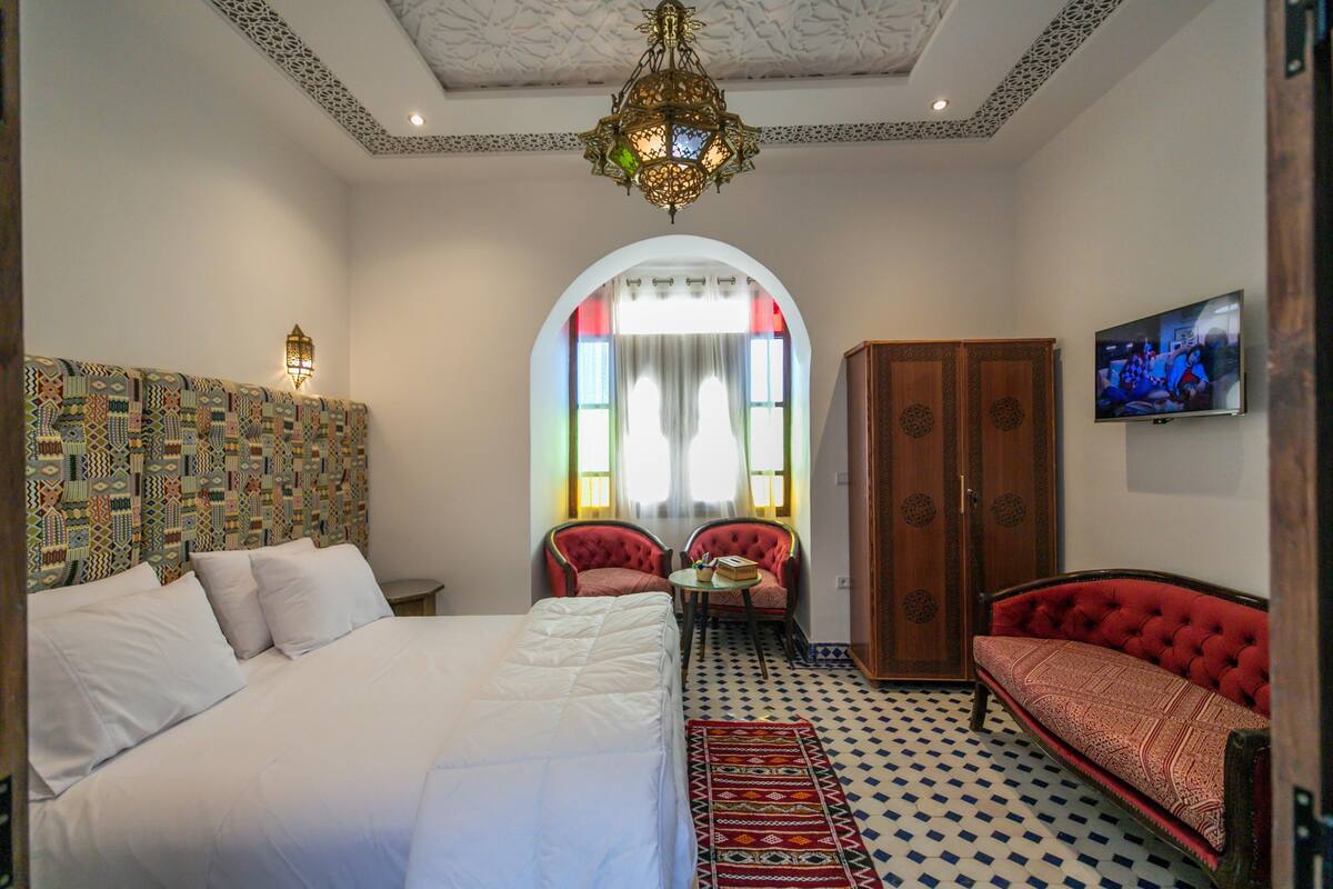 A bedroom with a large bed, two red armchairs, a wooden wardrobe, and a television on the wall.