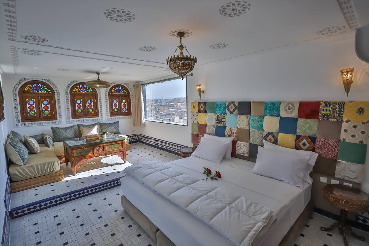 A hotel room with a large bed, a sofa, a wooden chest of drawers, a small table, and a view of the city through a large window.
