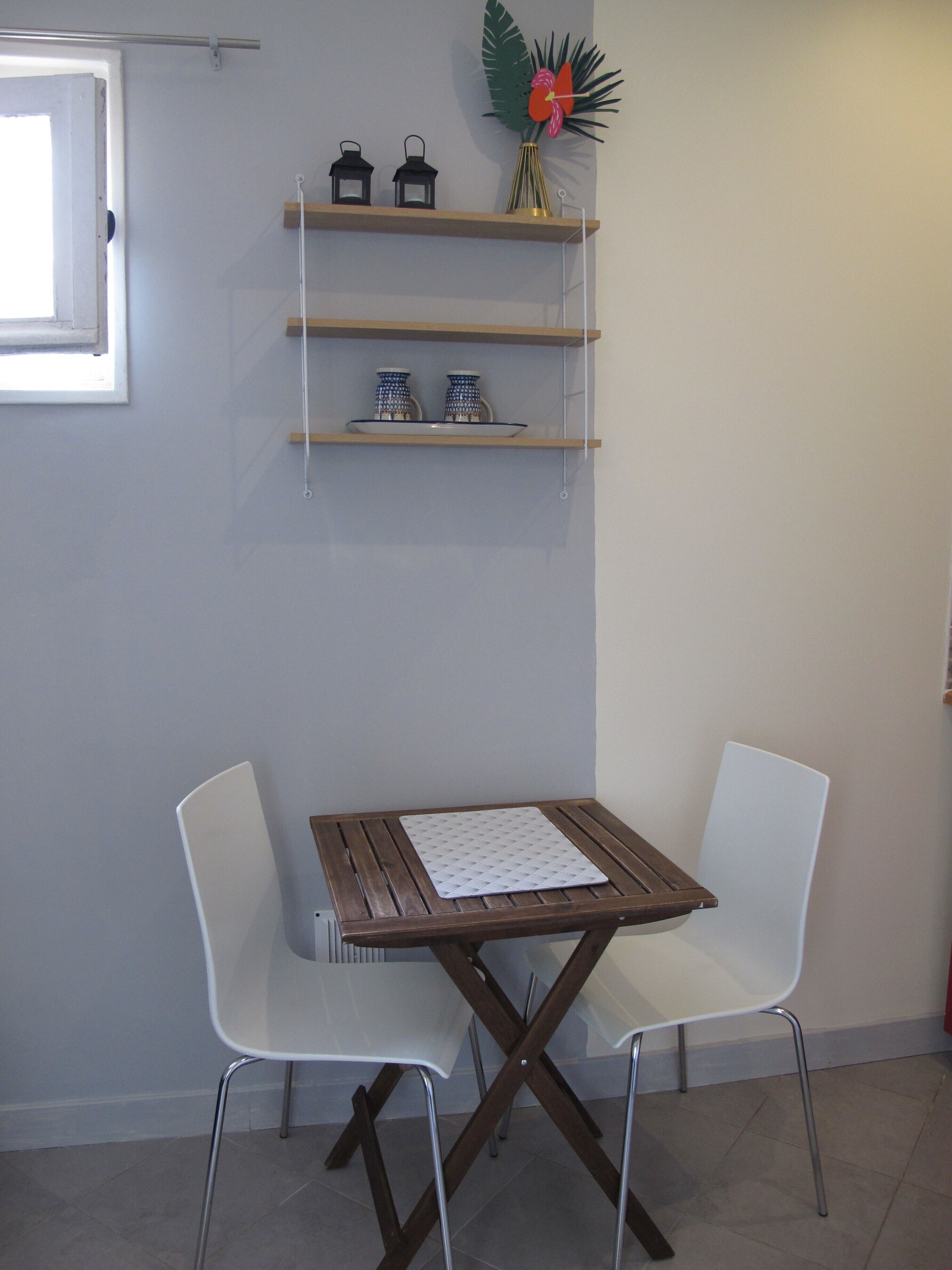 Een klein zithoekje met een houten tafel en twee witte stoelen. Daarboven bevinden zich drie planken, elk versierd met decoratieve objecten.