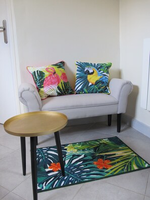 Ein Zimmer mit einem grauen Sofa, zwei farbenfrohen Kissen, einem kleinen runden Tisch und einem Teppich mit tropischem Blattmuster.