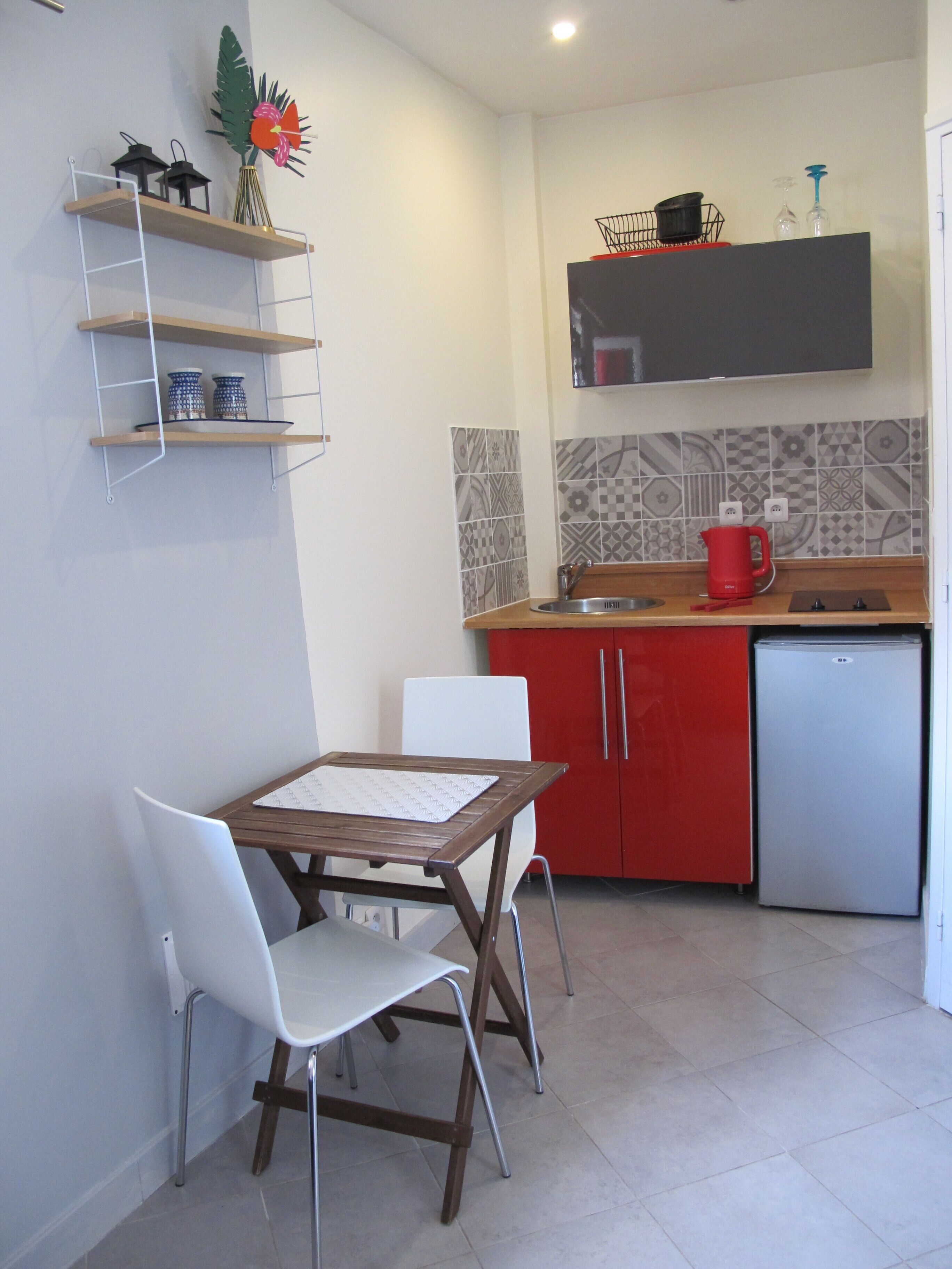 Een compacte keuken met een eethoek, bestaande uit een kleine tafel, stoelen, een koelkast en een plank met keukengerei.