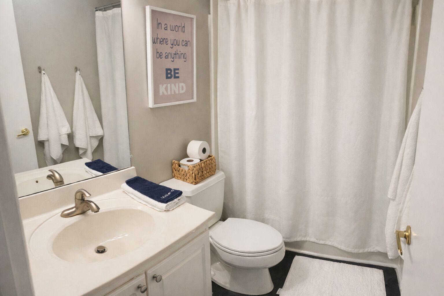 Et badeværelse med vask, toilet og bruseforhæng. Der er et indrammet citat på væggen og en kurv med toiletartikler på toilettet.