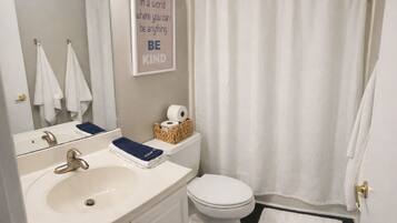 Et badeværelse med vask, toilet og bruseforhæng. Der er et indrammet citat på væggen og en kurv med toiletartikler på toilettet.
