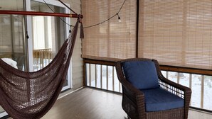 Une pièce avec un fauteuil en osier, un hamac et une vue dégagée sur l’extérieur grâce à de grandes fenêtres équipées de stores.