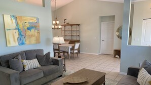 A living room with a gray sofa, a coffee table, and a dining area with a chandelier and a glass cabinet. - South Venice Hidden Gem (Venice)