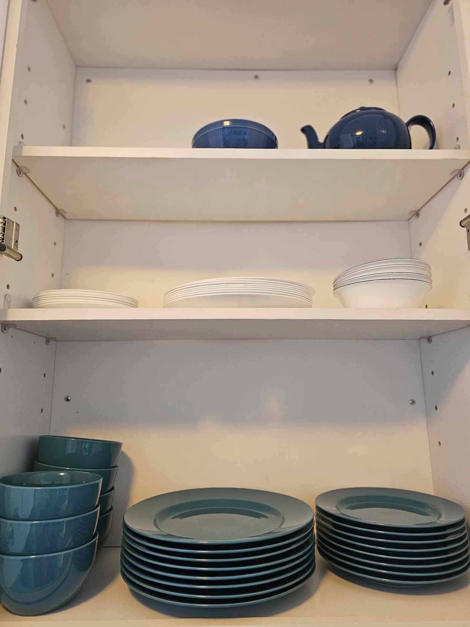 Una cocina con armarios y estantes blancos. Se observan pilas de platos blancos, cuencos azules y una tetera azul sobre los estantes.
