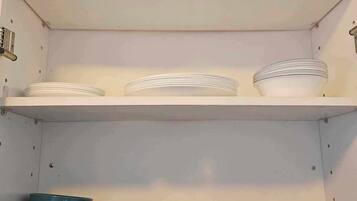 Una cocina con armarios y estantes blancos. Se observan pilas de platos blancos, cuencos azules y una tetera azul sobre los estantes.
