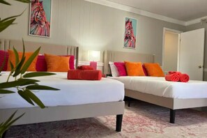 Ein Hotelzimmer mit zwei Betten, jeweils mit weißer Bettwäsche und orangefarbenen Kissen. An den Wänden befinden sich zwei Bilder, im Vordergrund ein Blumentopf und rechts eine Tür.