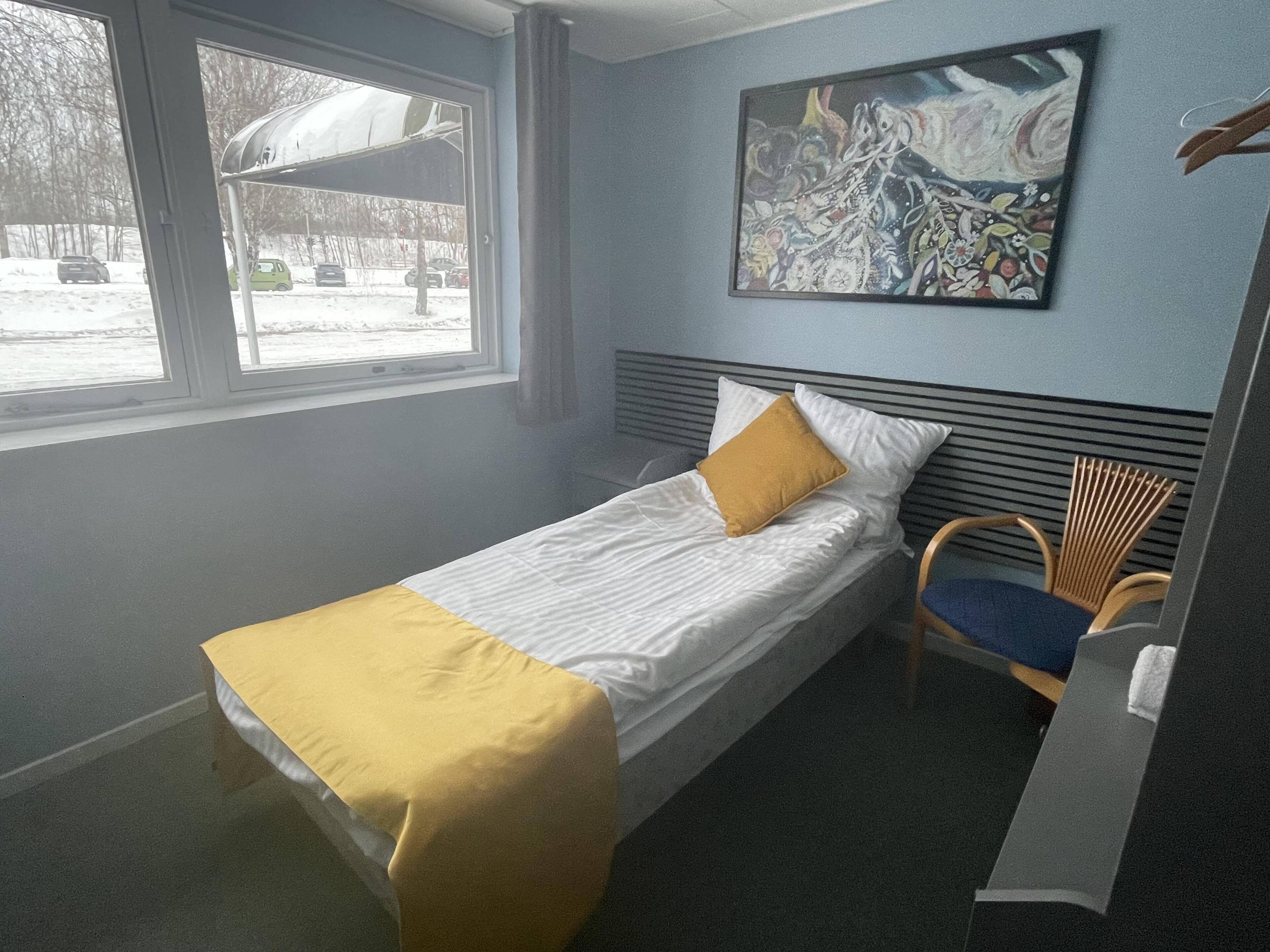 A single bed with a yellow blanket, a chair, a window with a view of parked cars and trees, and a painting on the wall.
