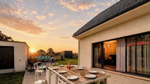 Un espace de restauration en plein air avec une table dressée pour quatre personnes, entourée de chaises, à proximité d’un bâtiment doté d’une grande porte-fenêtre coulissante.