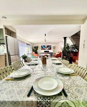Comedor con una mesa larga dispuesta para seis personas, rodeada de sillas, con una cocina y zona de estar al fondo.