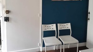 Une pièce avec une porte blanche, un mur bleu et un tableau représentant une scène de rue. Il y a deux chaises blanches dans la pièce.