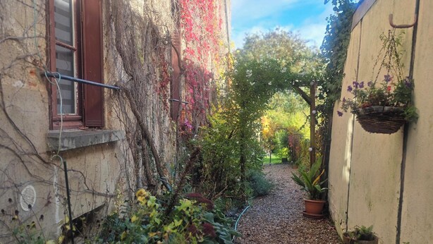 Une ruelle étroite avec un chemin de gravier, un bâtiment recouvert de lierre rouge et des jardinières suspendues.