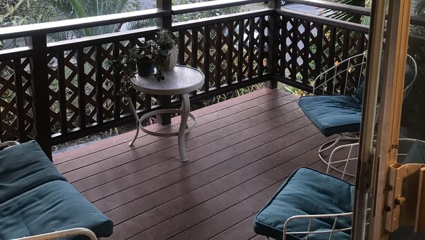 Ein überdachter Außenbereich mit Holzdeck, einem runden Tisch mit einem Topf und zwei blauen Sitzgelegenheiten mit Kissen.