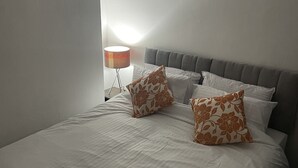 Una cama bien hecha con ropa de cama blanca y dos almohadas decorativas, una lámpara de noche y un cabecero gris.
