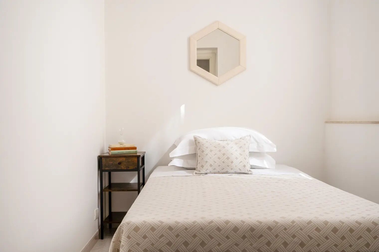 Quarto com cama, mesa de cabeceira com livros, espelho e colcha estampada.