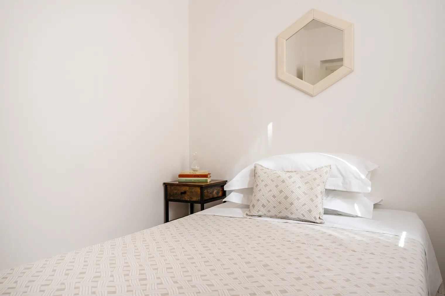 A bedroom with a bed, a bedside table with books and a lamp, and a wall-mounted mirror.