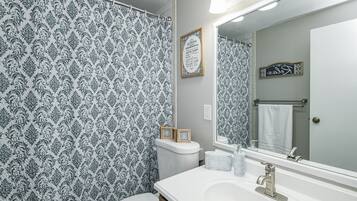 Een badkamer met een gordijn met een patroon, een lijst met een bord boven het toilet, een grote spiegel en een handdoekhouder.