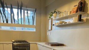 Cocina con estufa, horno y piso de madera. Se ve una ventana con plantas y un estante con utensilios de cocina.
