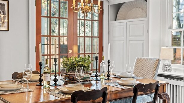 Une salle à manger avec une table en bois dressée pour quatre personnes, un lustre et une grande fenêtre donnant sur un paysage arboré.