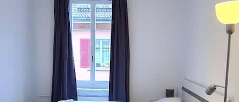 A single bed with a wooden floor, a bedside table, a lamp, and a window with curtains.