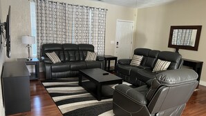 A modern living room with a black leather sofa, a black coffee table, and a striped rug. There is a ceiling fan, a window with patterned curtains, and a door. - Cozy Stay near UCHealth and Anschutz Hospitals  (Aurora)