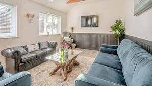 Ein Wohnzimmer mit Glastisch, grauem Sofa, blauem Sofa, Holzbalken an der Decke, Wand-Fernseher und Wanddeko mit Elefantenkopf.