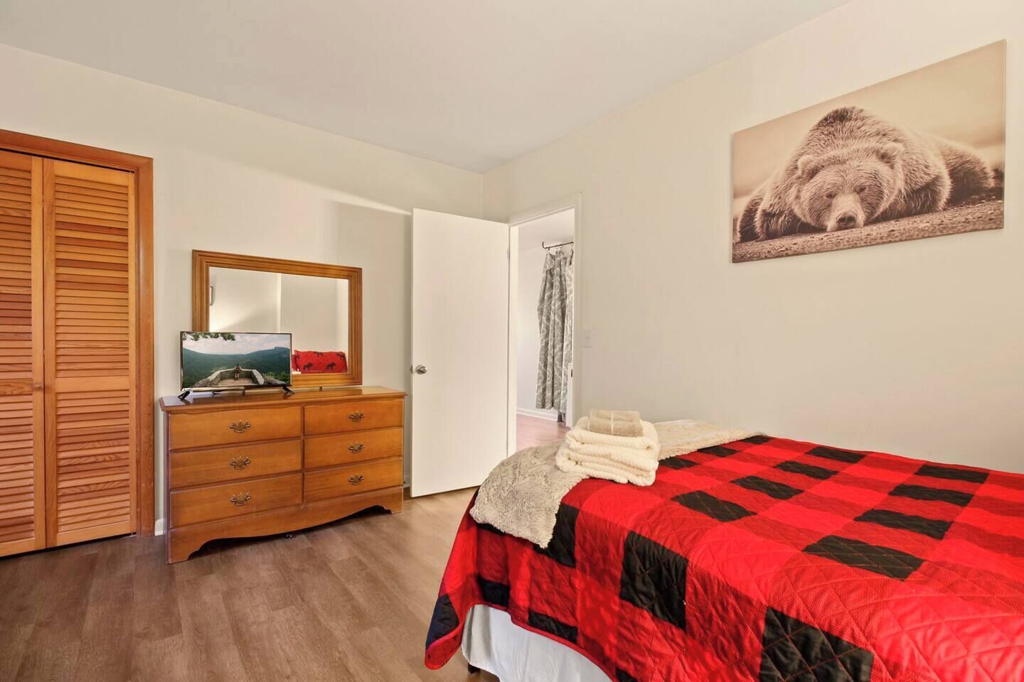 A bedroom with a wooden dresser, a bed with a red and black quilt, a mirror, and a bear picture on the wall.