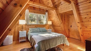 Un dormitorio acogedor en el ático, con techo inclinado, una cama con colcha verde, dos mesitas de noche con lámparas y una ventana con cortinas.