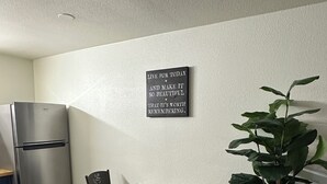 A compact kitchen with a built-in refrigerator, a dining table with two chairs, a potted plant, and a motivational quote on the wall.