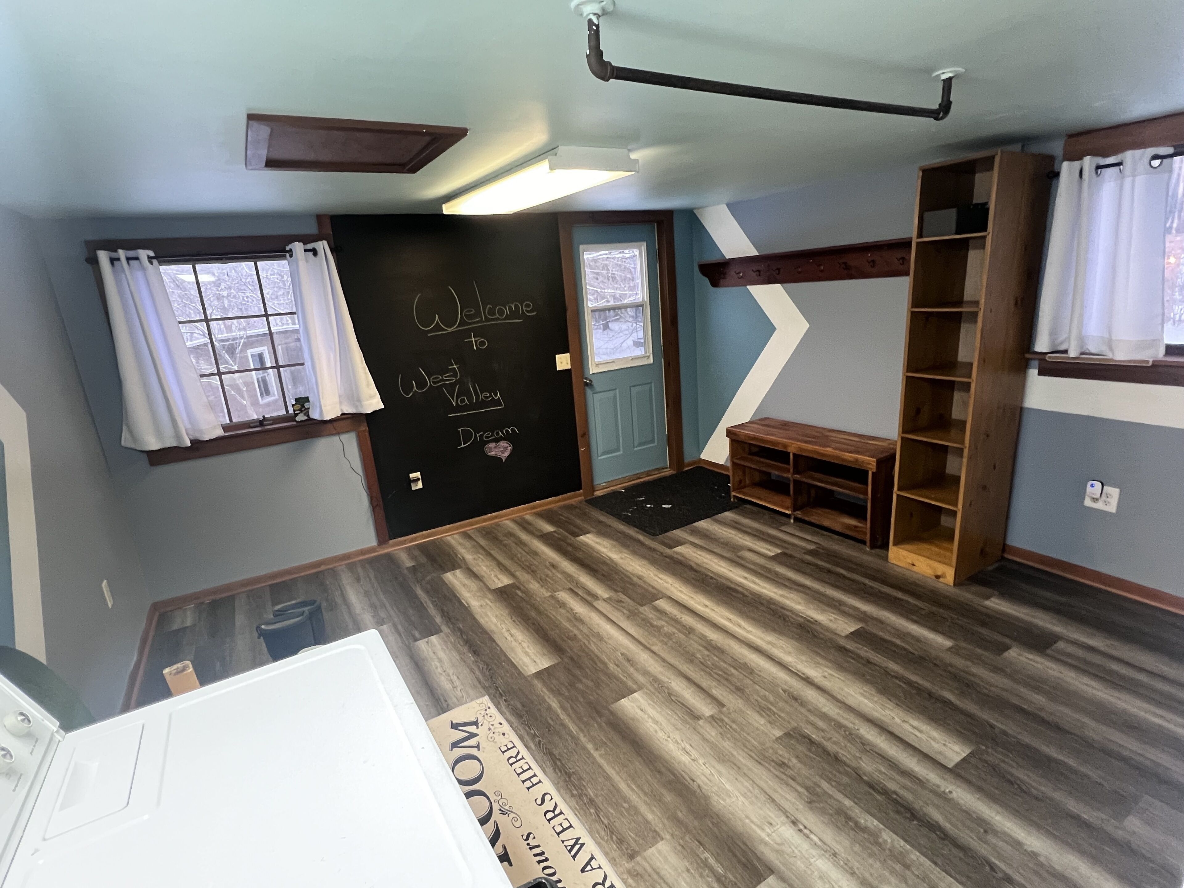 A room with a wooden floor, a chalkboard with 'Welcome to West Valley Dream' written on it, a wooden bench, and a bookshelf.