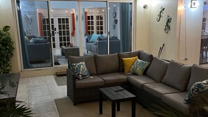 Un salon comprenant un canapé d’angle, une table basse et un ventilateur de plafond. Une porte-fenêtre coulissante donne accès à un espace extérieur aménagé avec un ensemble de mobilier de jardin.