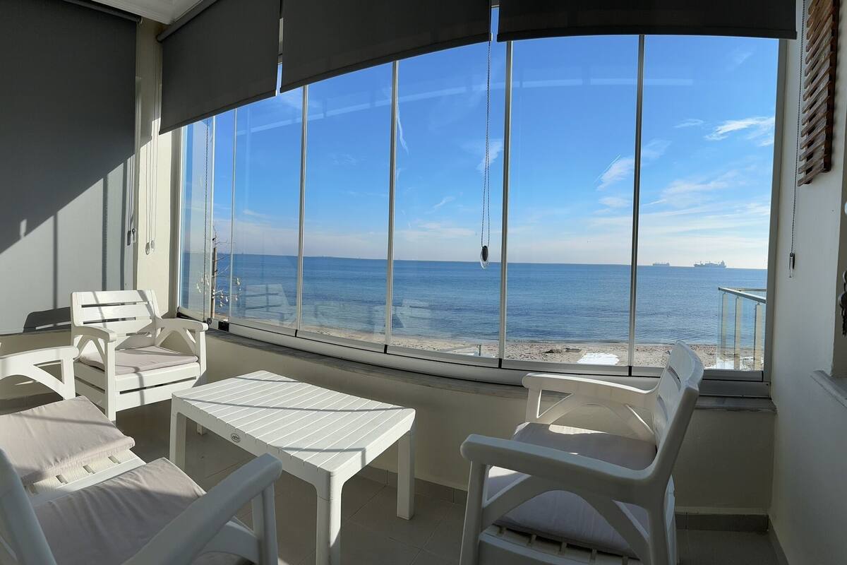 A balcony with white furniture, a table, and a view of the sea and ships.