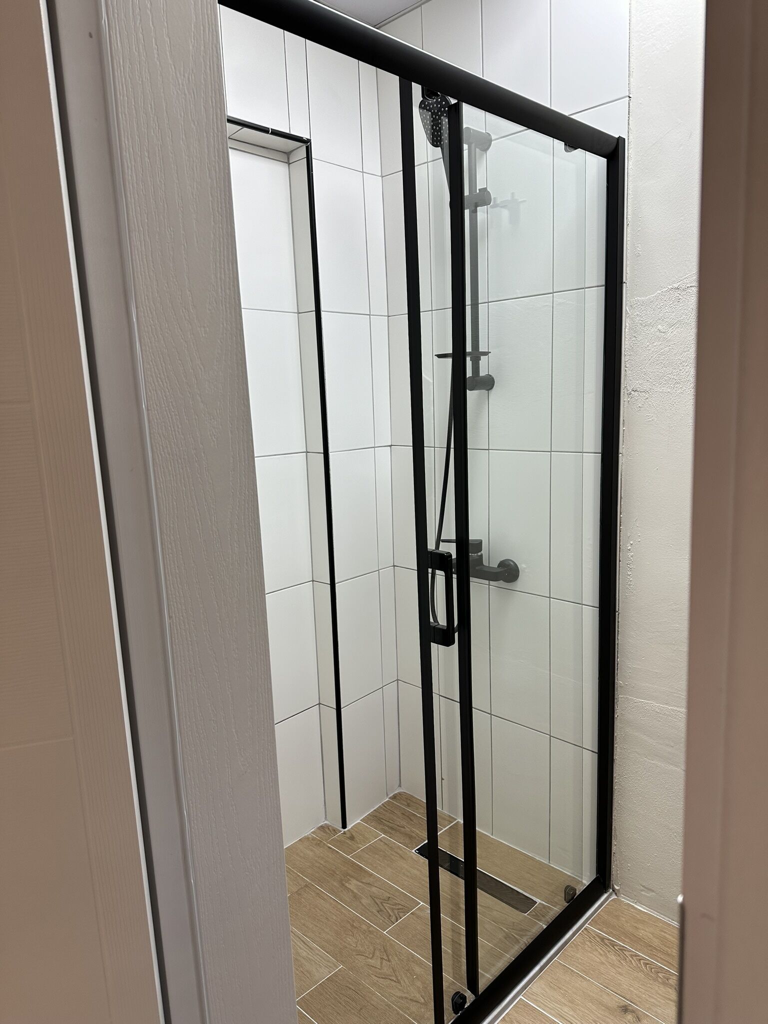 A glass-enclosed shower with a black frame, white tiled walls, and a wooden floor.