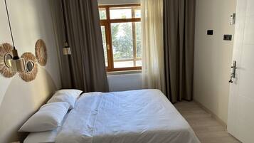 Una cama bien hecha con ropa blanca en una habitación con un ventanal grande, cortinas y una lámpara fijada a la pared.