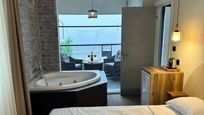 A hotel room with a large bed, a jacuzzi, a wooden nightstand, a table with two chairs, and a balcony with a view of the sea. - Kingway Suite (Tekirdag)