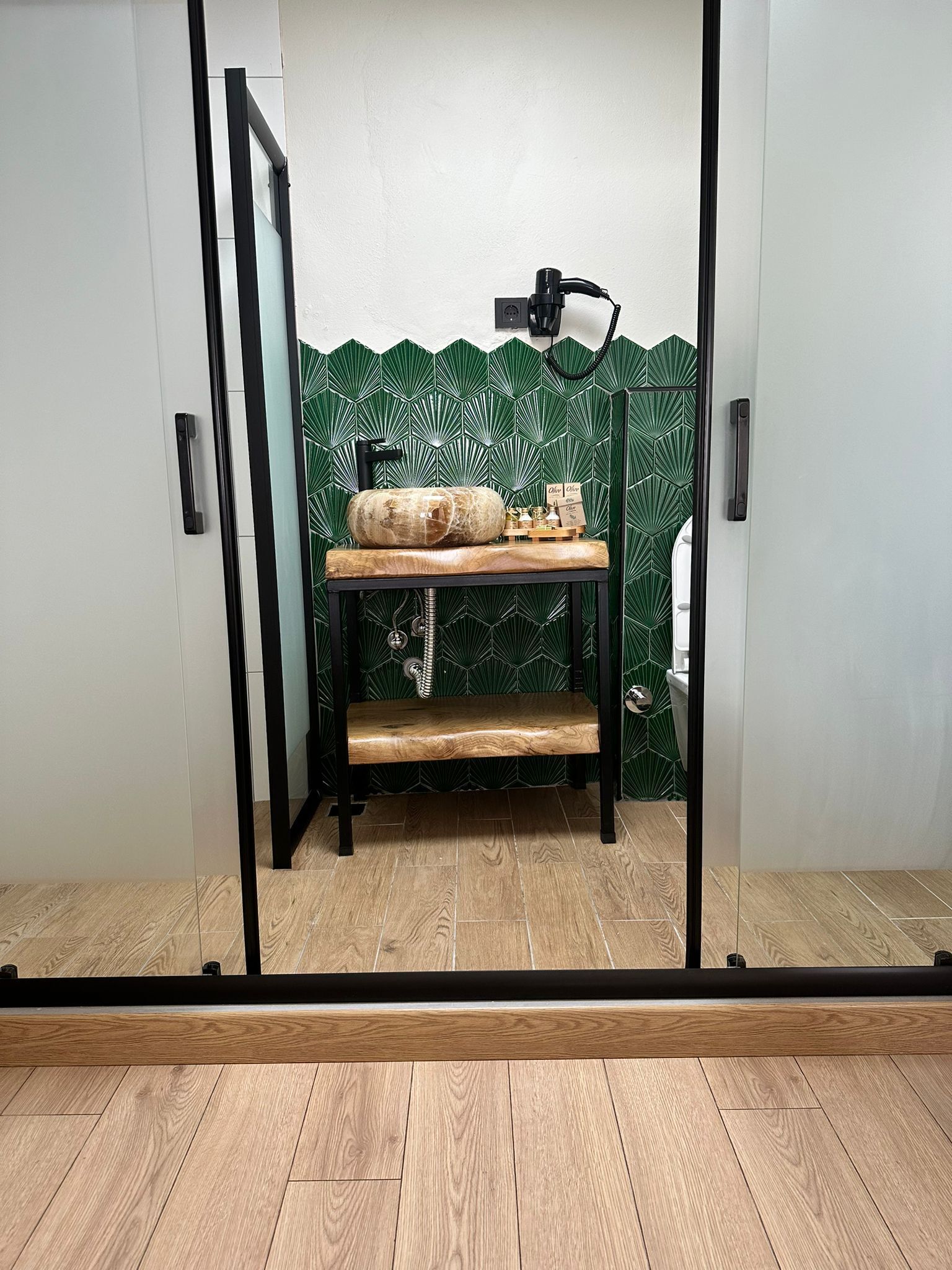 Une pièce avec un banc en bois, une étagère fixée au mur où sont rangées des bouteilles, et un mur décoré d’un motif de feuilles vertes.