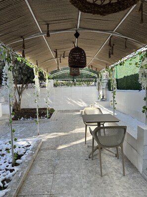 An outdoor patio area with tiled flooring, covered by a thatched roof, featuring tables and chairs, surrounded by greenery and white flowering plants. - Kingway Suite (Tekirdag)