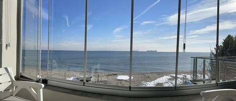 A view from a covered outdoor area with a glass wall, overlooking a beach with snow patches and a calm sea.