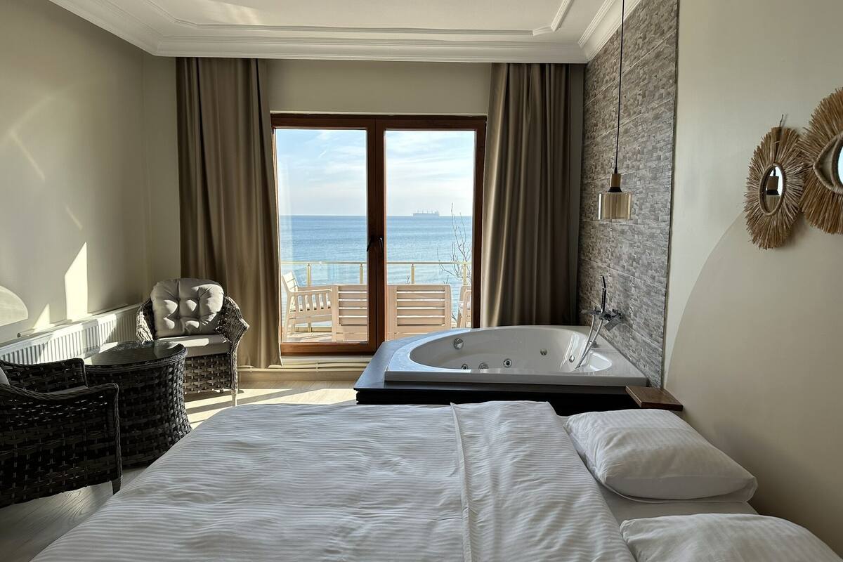 A hotel room with a large bed, a bathroom with a bathtub, a wicker chair, a view of the sea, and a balcony.